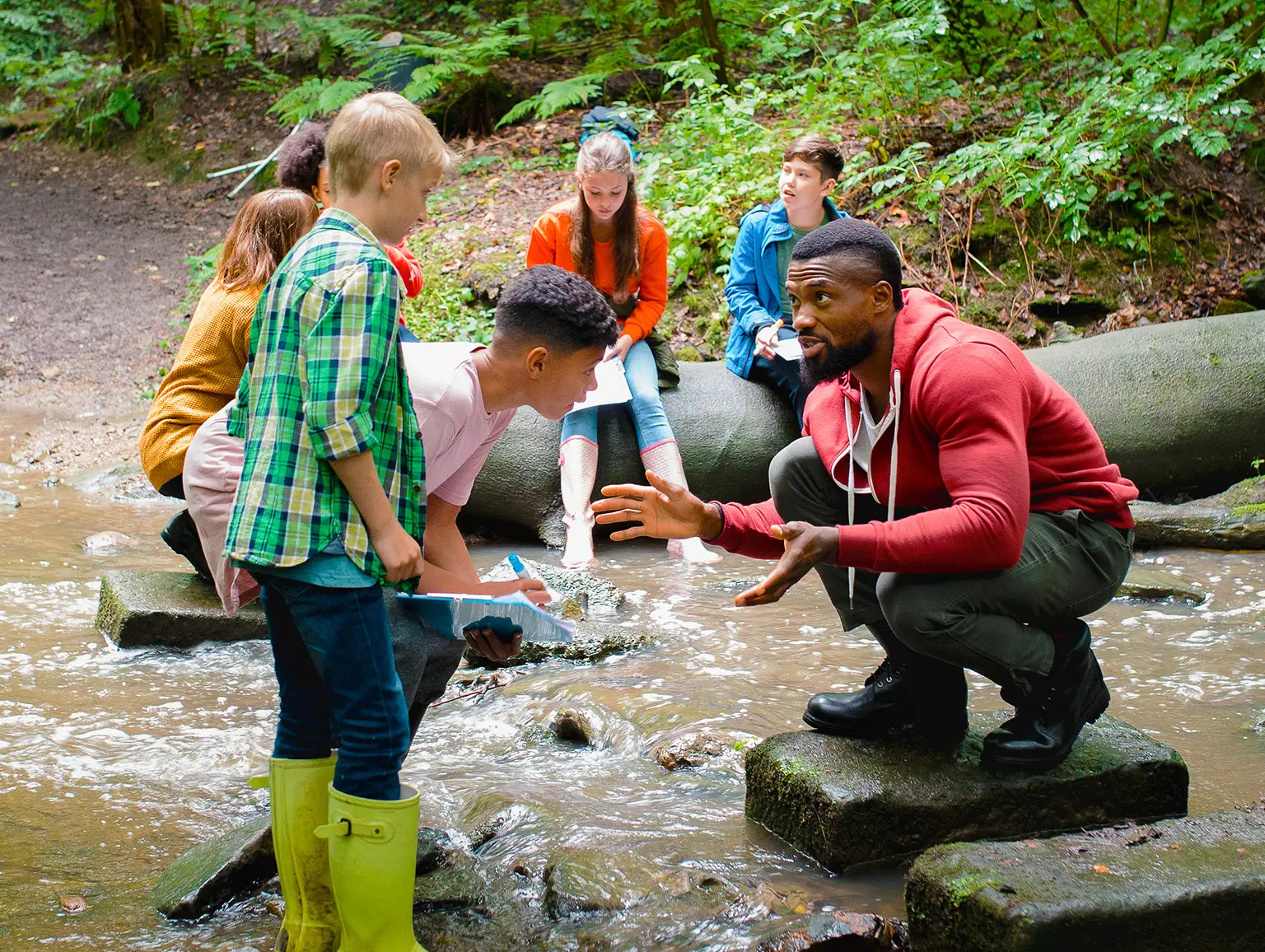 An adult helping a group of children during an outdoor activity by a creek.