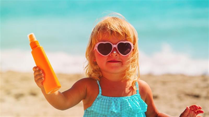 Little girl in sunglasses on a beach holding up a bottle of sunscreen
