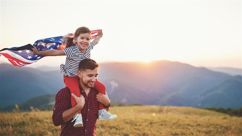Dad with daughter on his shoulders wearing red, white, and blue and an American flag billowing out behind her