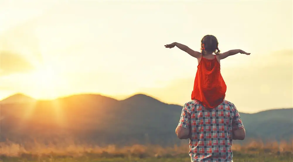 A little girl on dad's shoulders with arms spread out in the sunrise