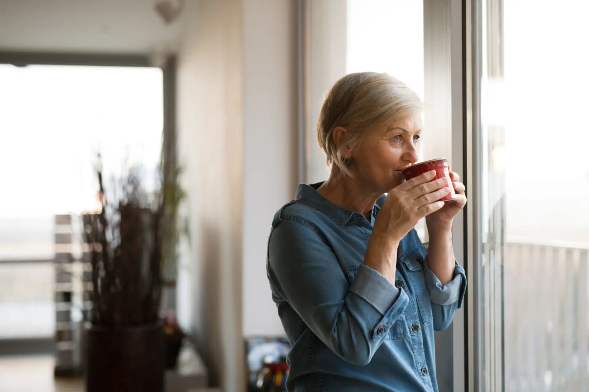 An older woman looking sipping from a coffee mug looking out the window