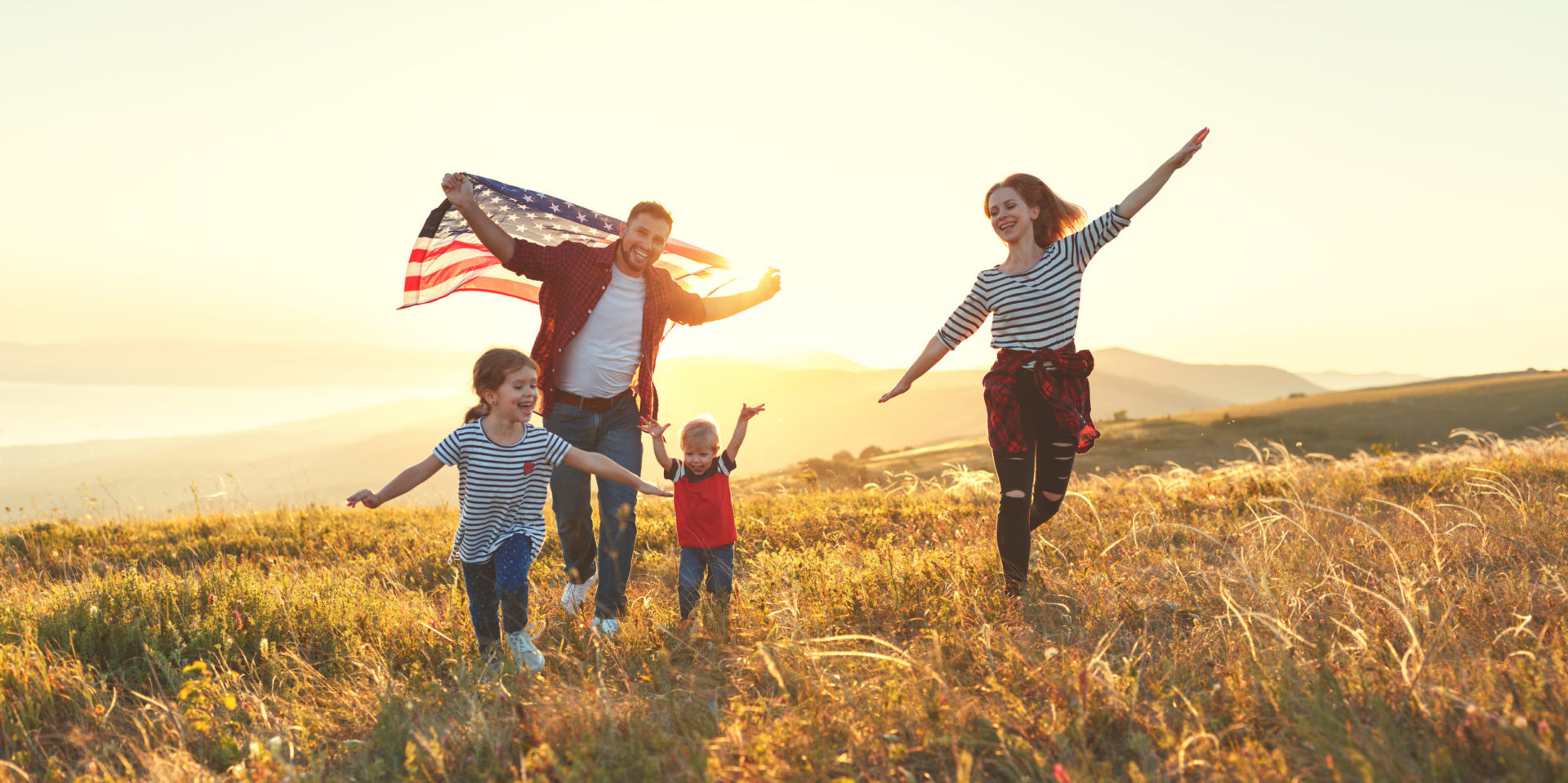 Familia de cuatro con bandera americana corriendo por un campo dorado