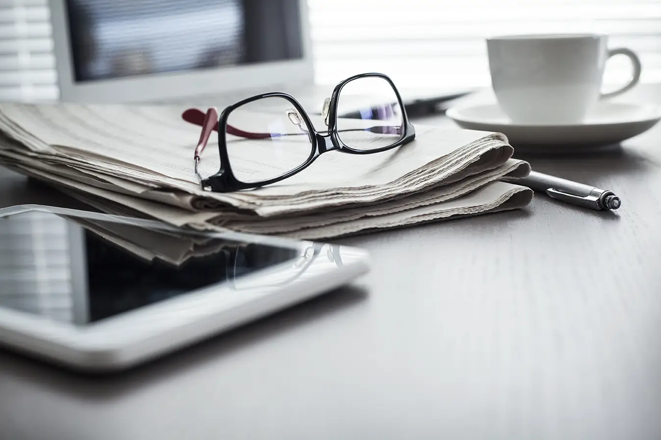 Eyeglasses on top of a newspaper next to a coffee cup and a tablet on a table