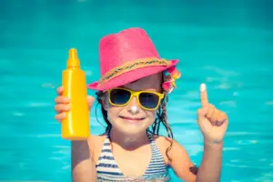 A little girl with a hat on in the pool holding sunscreen in her hand