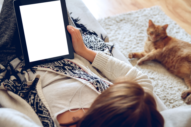 Woman stretched out on the couch holding a tablet with cat look up at her