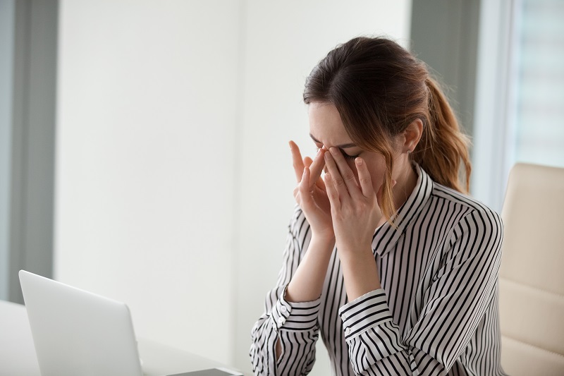 Mujer en la oficina frotándose los ojos mientras está sentada frente a su portátil abierto