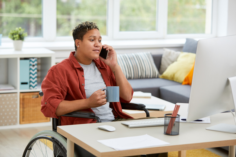 Persona sentada en una silla de ruedas en el escritorio de su casa mientras habla por teléfono con una taza de café en la mano