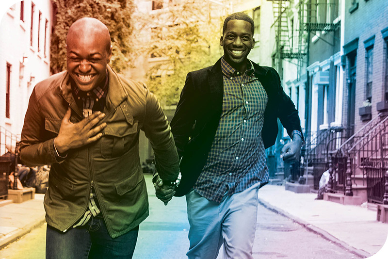 Dos hombres riendo y cogidos de la mano mientras caminan juntos por la calle, con un arco iris superpuesto
