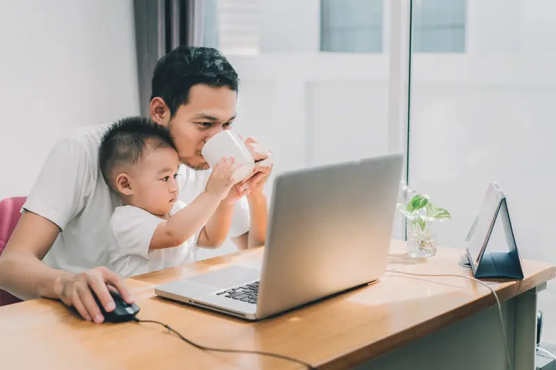 A father drinking coffee at his computer with his son on his lap holding the coffee cup