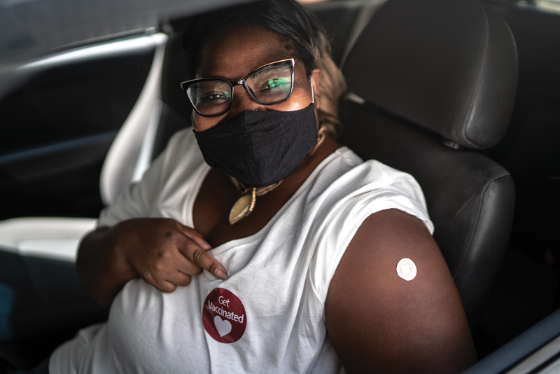 Woman in car point to her Get Vaccinated sticker after receiving her vaccine