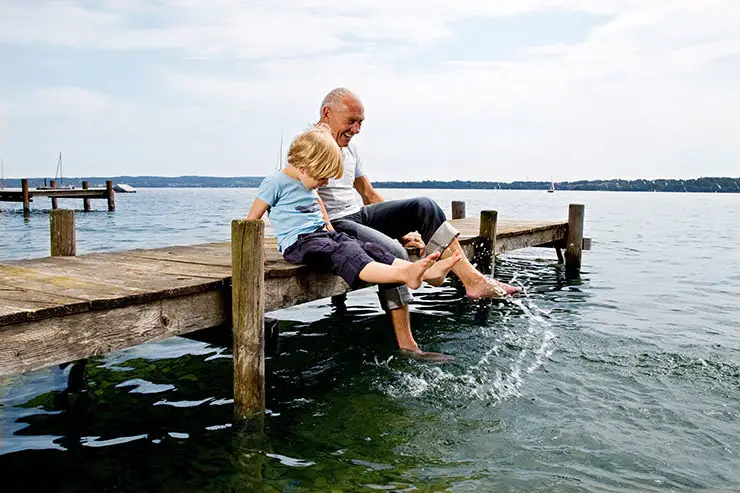 A boy and an older man sitting on a doc splashing with their feet in the water