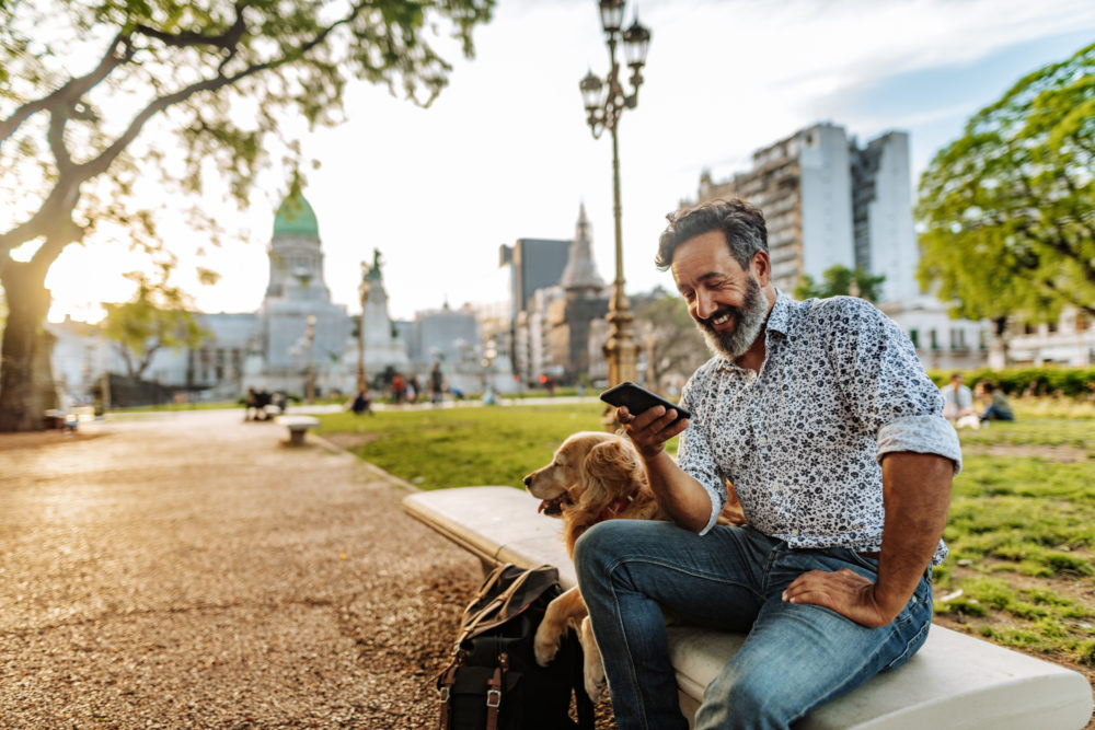 Hombre usando un teléfono inteligente en un banco con su perro en Buenos Aires