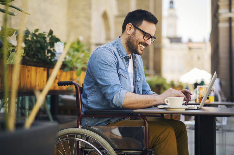 Joven guapo en silla de ruedas sentado en una cafetería al aire libre trabajando con un ordenador portátil