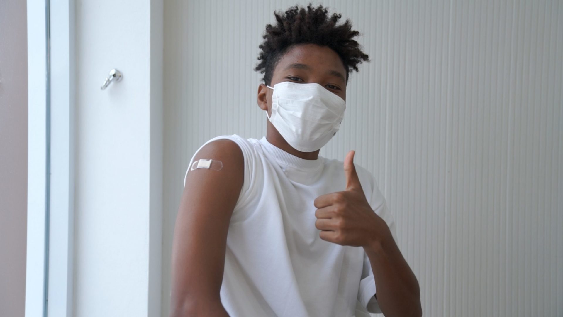 Boy wearing a face mask giving thumbs up with a bandaid on his arm after getting his COVID-19 vaccine