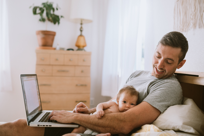 Un padre trabaja con su portátil en la cama con un bebé en brazos