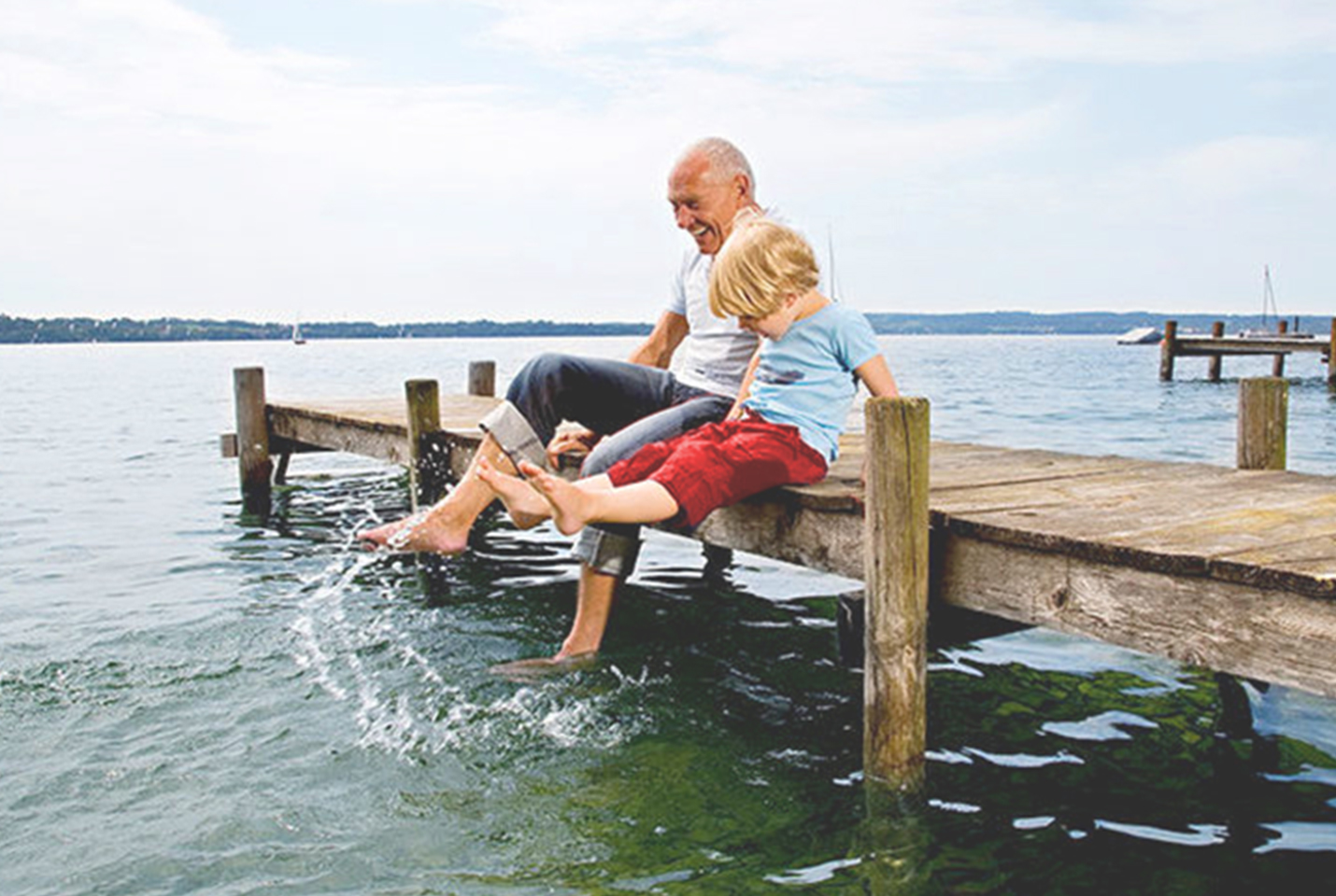 A grandfather and young boy splashing their feet on a dock at a lake