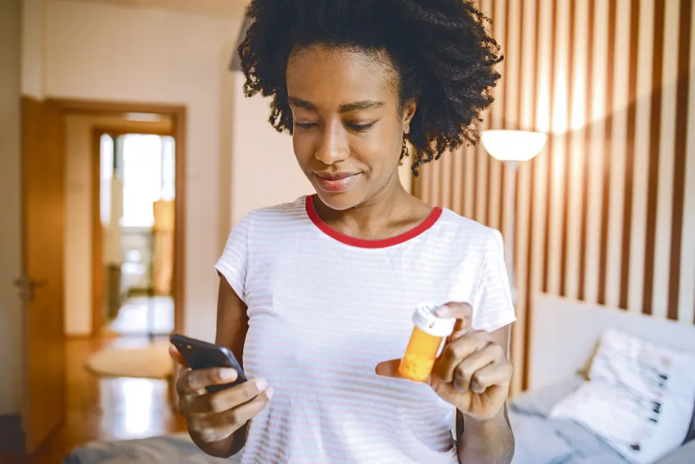 Una mujer sostiene un frasco de pastillas en una mano y utiliza un teléfono inteligente en la otra en su casa