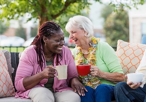 Two senior women sitting at an outdoor patio holding coffee mugs laughing