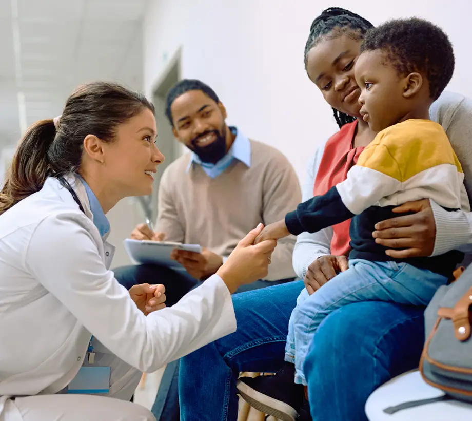 Doctor crouching to engage with a young boy on his parent's lap at a clinic.