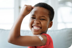 A young boy in a red shirt smiles and flexes his arm to show the bandaid he got after getting a shot