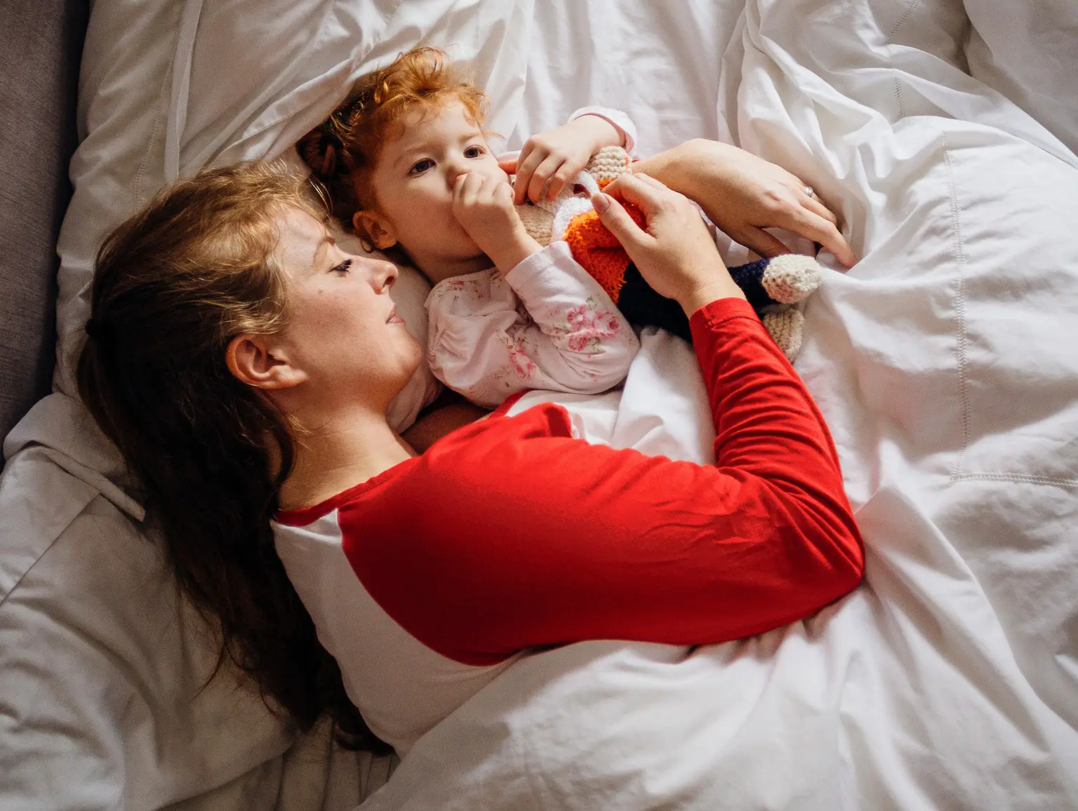 A parent and child lying in bed. The child is sucking their thumb and holding a stuffed animal.