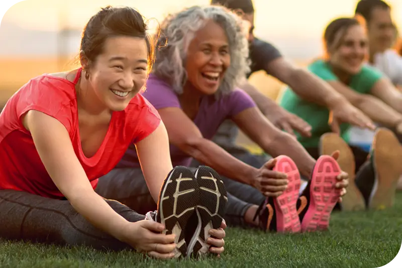 Two persons stretch during an outdoor fitness activity. They are wearing sportswear and smiling.