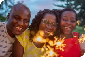 Two adults and a child smile as they look at a sparkler on what appears to be a summer evening.