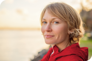 A person with a soft smile looks into the camera lense as they stand in front of a lake background