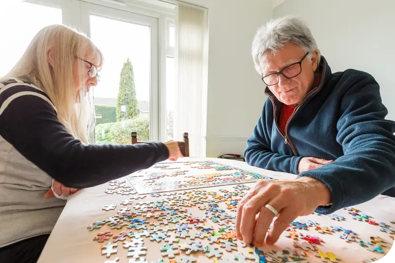 Two persons complete a puzzle in a room with a lot of natural light.