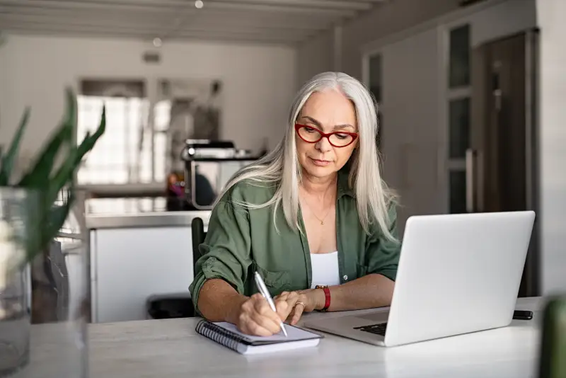 A senior woman takes notes while sitting in front of her laptop.