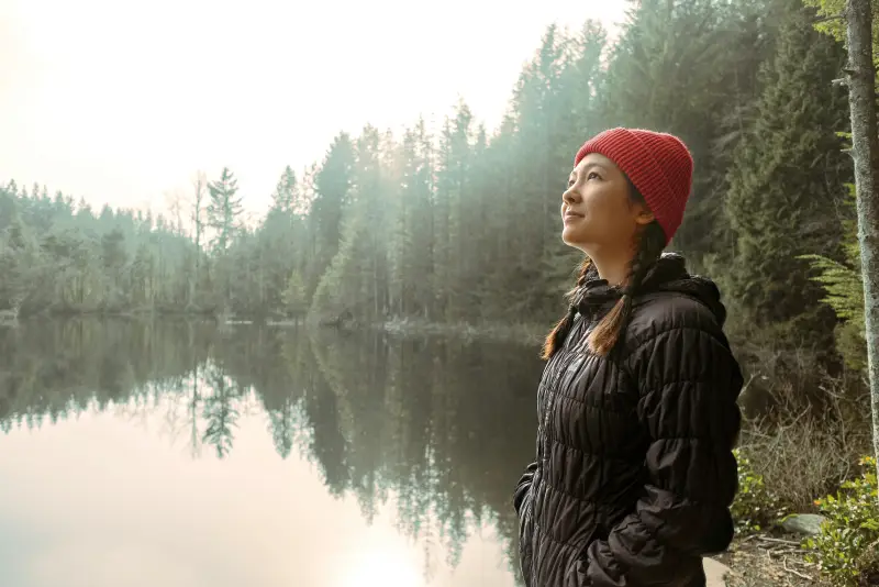 Young woman starring peacefully at the sky while standing by a lake bordered with pine trees