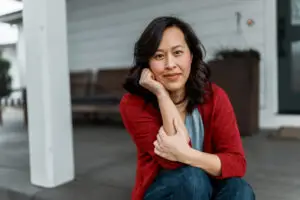 Portrait of a woman with a soft smile looking directly at the camera while sitting on the step leading to the covered patio and front door of her home.