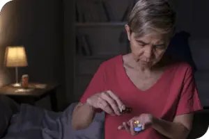 A person takes a pill from an open pill bottle, with a dimly lit bedroom visible in the background at night.