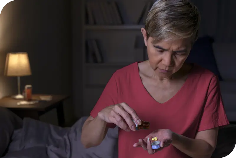 Una persona toma una pastilla de un frasco abierto, con un dormitorio poco iluminado visible al fondo por la noche.