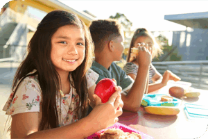 Three children sit at an outdoor table as one smiles at the camera holding an apple