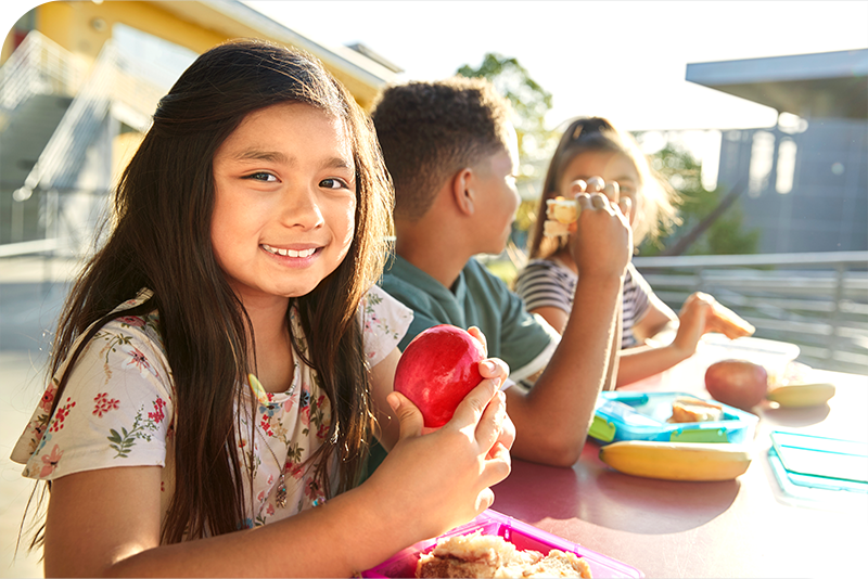 Three children sit at an outdoor table as one smiles at the camera holding an apple