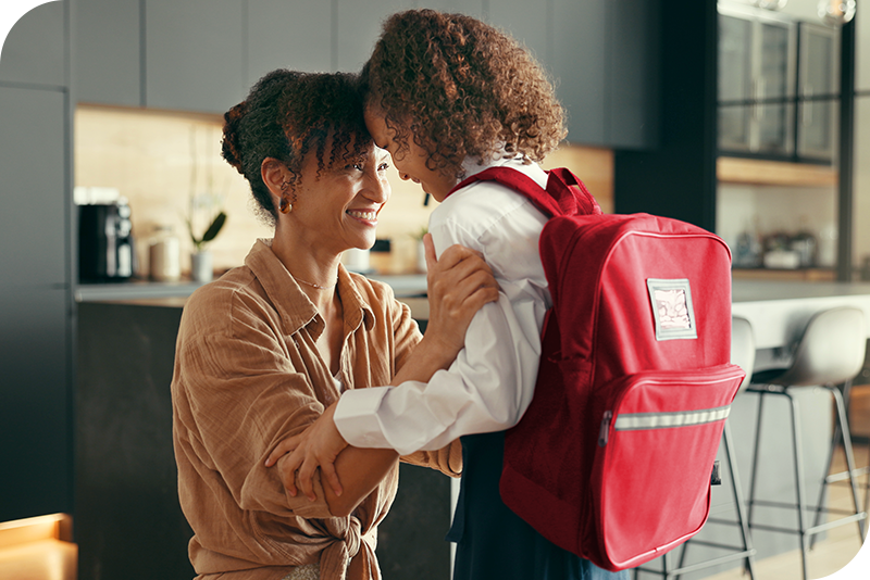Mother smiling while buttoning her child’s shirt as the child wears a backpack