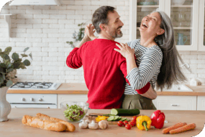A couple dances in the kitchen in front of a cutting board covered in vegetables