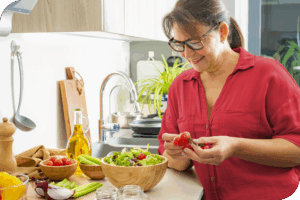 A woman smiles as she adds tomatoes to a salad in a brightly lit kitchen