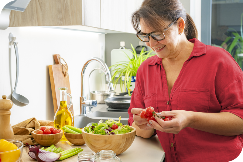 A woman smiles as she adds tomatoes to a salad in a brightly lit kitchen