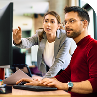 Two persons sit in front of a computer as one of them points at it