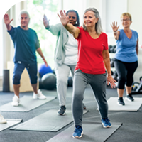 A group of people practice aerobics in a gym lit by natural light coming in through a large window
