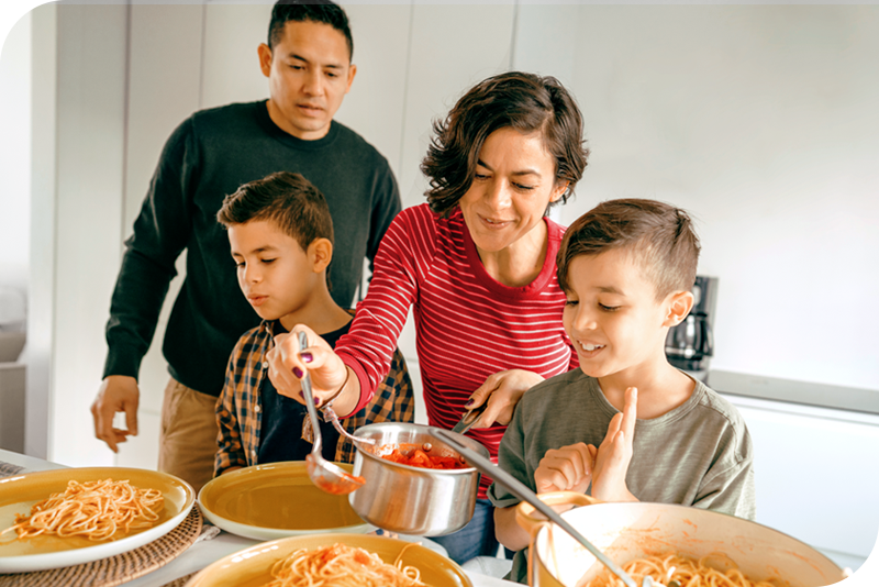 A family of four smiles as the mother serves plates of spaghetti
