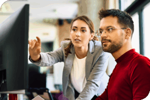 Two persons in an office setting look at a monitor as one of them points at it and seems to be saying something