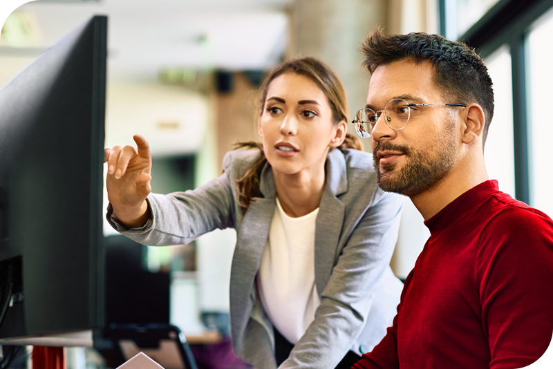Two persons in an office setting look at a monitor as one of them points at it and seems to be saying something