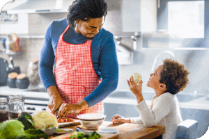 An adult in a red apron smiles and looks down at a child as they appear to be cooking in a brightly lit kitchen