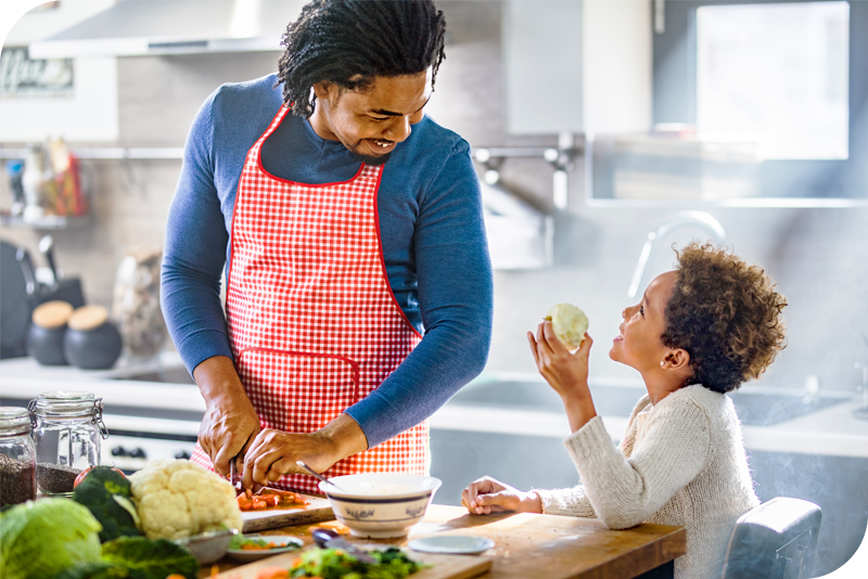 An adult in a red apron smiles and looks down at a child as they appear to be cooking in a brightly lit kitchen
