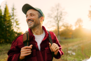 A man in a red shirt and a hat smiles as a holds on to his backpack straps, in front of a sunset-lit fall background