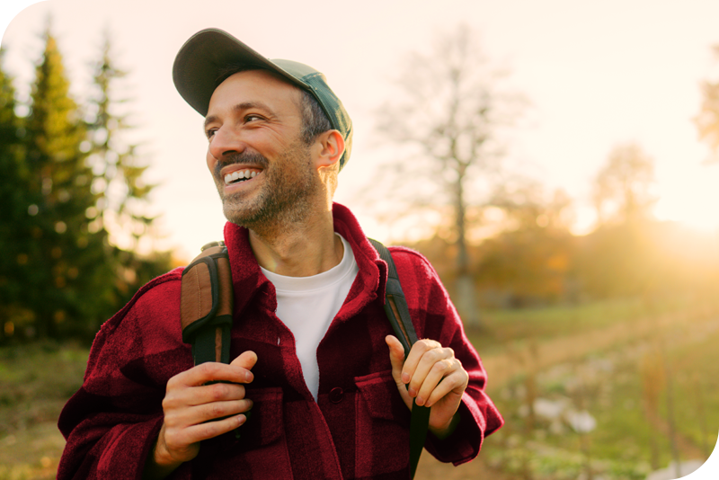 A man in a red shirt and a hat smiles as a holds on to his backpack straps, in front of a sunset-lit fall background