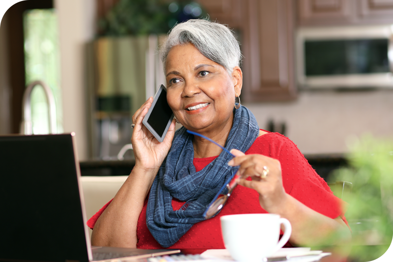 A woman sits at a table in her home, in front of a laptop, as she smiles and holds a phone to her ear